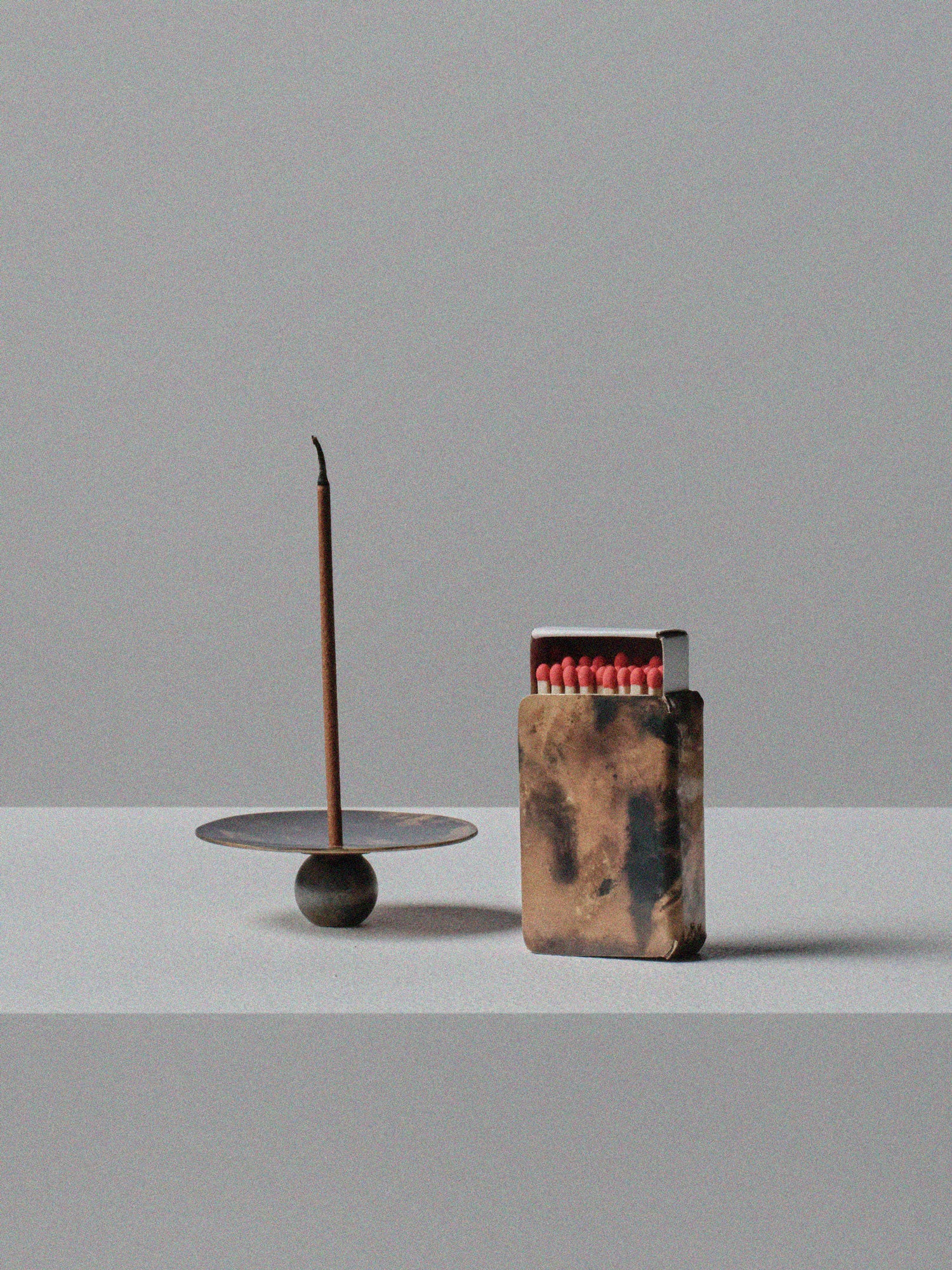 Handmade brass incense holder with burning stick and patinated brass matchbox cover by 13 Knives, displayed on a minimal grey surface