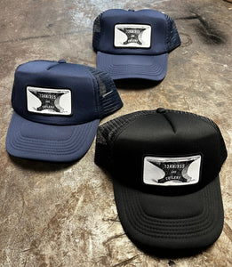 Three trucker hats in navy and black with a 13knives and cutlery anvil logo on a metal shop workbench 